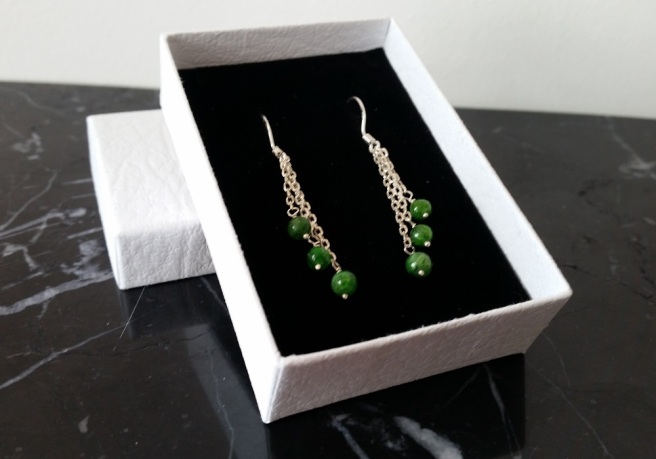 A pair of green and silver earrings in a white box on a black marble background